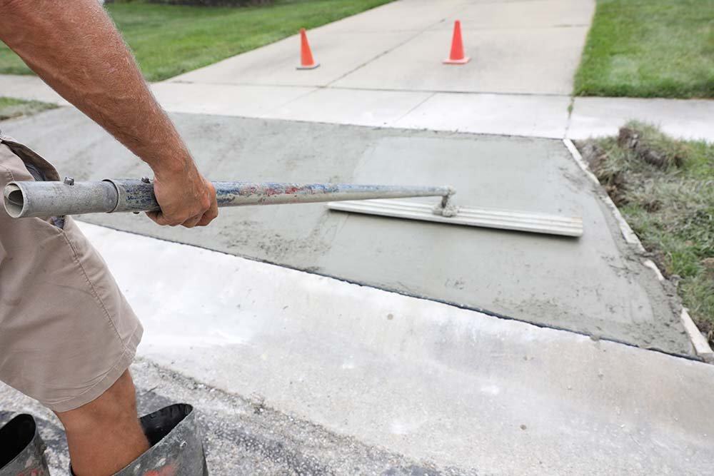 Pouring and smoothing concrete on a sidewalk with a trowel, with traffic cones in the background.